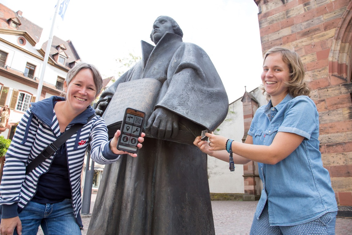 Jugendreferentin Constanze Harant (r.) und ihre katholische Kollegin zeigen den QR-Code im Ärmel der Luther-Statue in Landau. Er ist einer der Stationen, an die die App Actionbound for Future Interessierte führt.