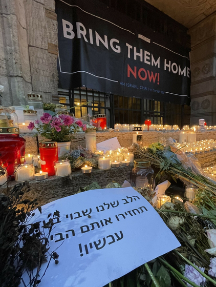 Nie wieder ist jetzt - Demonstration gegen Antisemitismus. Im Vordergrund sind Blumen und Kerzenhalter mit brennenden Kerzen auf einer Treppe aufgestellt. Dahinter ist ein Transparent mit dem Spruch "Bring the home now" an einem Gitter angebracht.