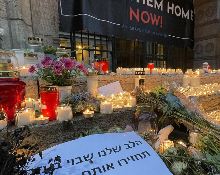 Nie wieder ist jetzt - Demonstration gegen Antisemitismus. Im Vordergrund sind Blumen und Kerzenhalter mit brennenden Kerzen auf einer Treppe aufgestellt. Dahinter ist ein Transparent mit dem Spruch "Bring the home now" an einem Gitter angebracht.