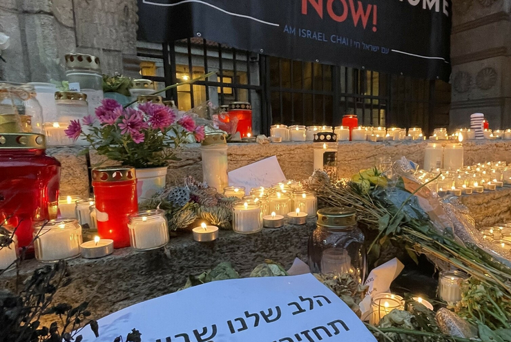 Nie wieder ist jetzt - Demonstration gegen Antisemitismus. Im Vordergrund sind Blumen und Kerzenhalter mit brennenden Kerzen auf einer Treppe aufgestellt. Dahinter ist ein Transparent mit dem Spruch "Bring the home now" an einem Gitter angebracht.