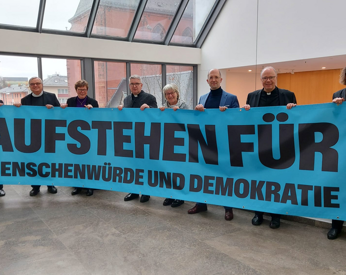 Eine Gruppe Männer und Frauen stehen nebeneinander hinter einem Transparent mit der Aufschrift "Aufstehen für"