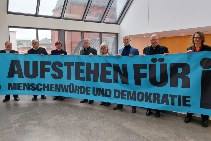 Eine Gruppe Männer und Frauen stehen nebeneinander hinter einem Transparent mit der Aufschrift "Aufstehen für"
