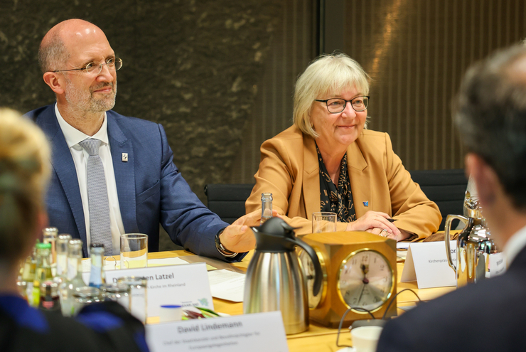 Ein mann und eine Frau sitzen an einem großen Konferenztisch, andere Menschen sind von hinten auf dem Bild zu sehen.
