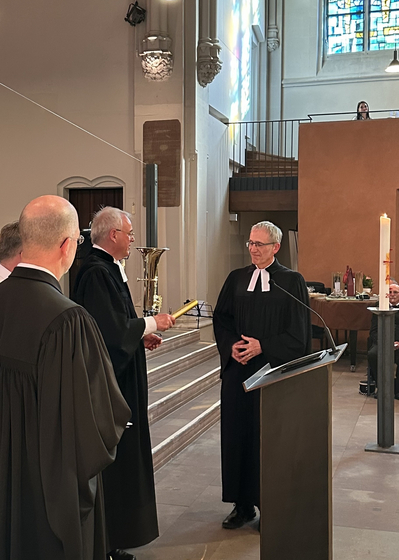 1 Mann im Talar übergibt einen goldenen Staffelstab an einen anderen Mann im Talar