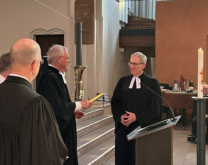 1 Mann im Talar übergibt einen goldenen Staffelstab an einen anderen Mann im Talar