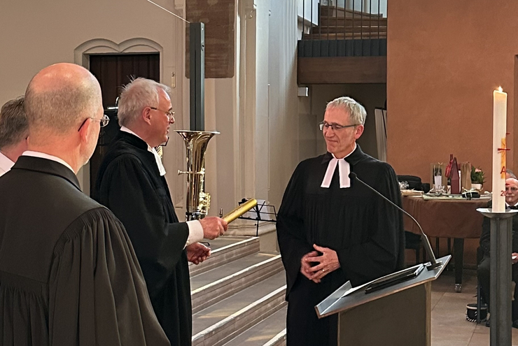 1 Mann im Talar übergibt einen goldenen Staffelstab an einen anderen Mann im Talar