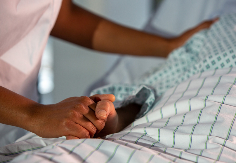Eine Person liegt auf einem Bett. Ihre Hand wird von einer anderen Person gehalten. Auf dem Bild sind nur die Hände zu sehen.