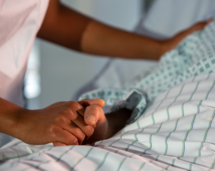 Eine Person liegt auf einem Bett. Ihre Hand wird von einer anderen Person gehalten. Auf dem Bild sind nur die Hände zu sehen.