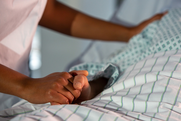 Eine Person liegt auf einem Bett. Ihre Hand wird von einer anderen Person gehalten. Auf dem Bild sind nur die Hände zu sehen.