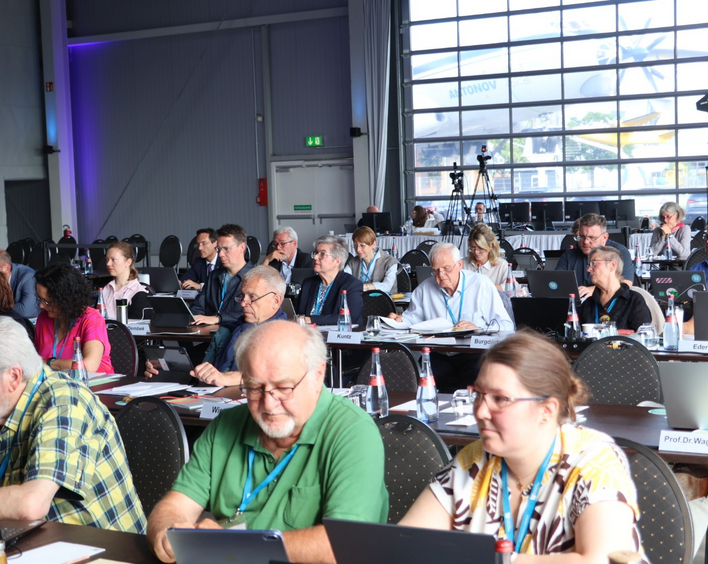 Gruppe MEnschen in einem Saal sitzt in Reihen an Tischen.