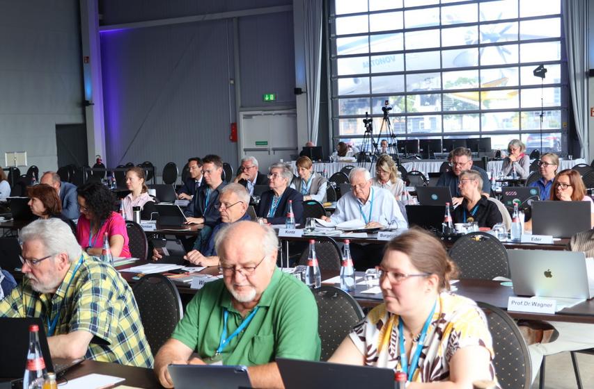 Gruppe MEnschen in einem Saal sitzt in Reihen an Tischen.