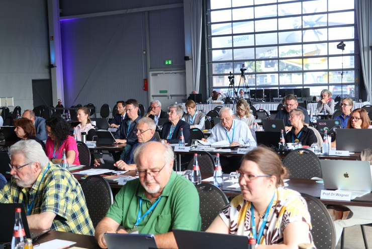 Gruppe MEnschen in einem Saal sitzt in Reihen an Tischen.